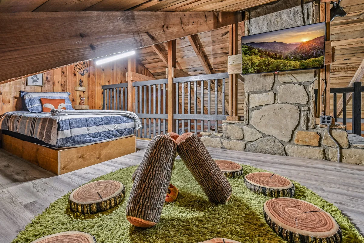 A cozy lodge living area with a striped bed, a rustic stone fireplace, a TV positioned to display scenic mountain views, and playful wooden log seating. Large windows allow natural light to spill in, connecting the interior to the outdoors and providing a comforting, welcoming space for guests and their pets.