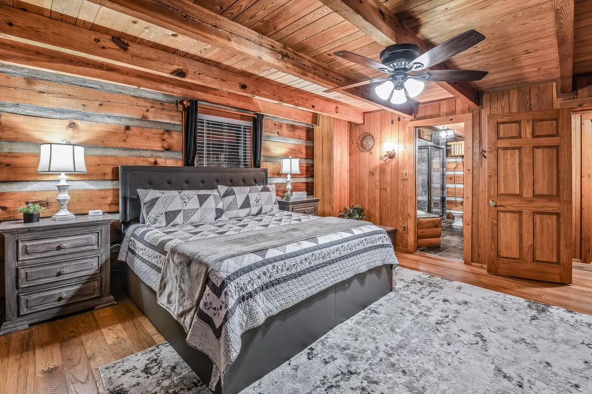 A tranquil bedroom with a bed covered in a patterned quilt, wooden nightstands with lamps on either side, and a textured area rug. The space is framed by a wooden ceiling and walls that emit a cozy, rustic charm. Soft morning light streams through an unseen window, creating an inviting atmosphere ideal for rest.