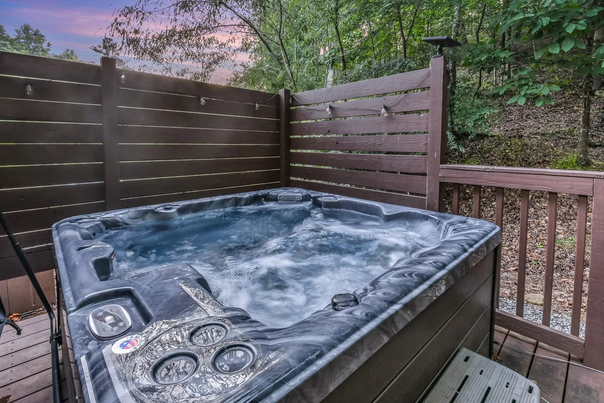 A private outdoor hot tub sits on a wooden deck surrounded by lush greenery. The inviting bubbling water is positioned for relaxation, with ample space for guests to enjoy the fresh mountain air and peaceful views. Ambient light hints at the possibility of evening dips under the stars.