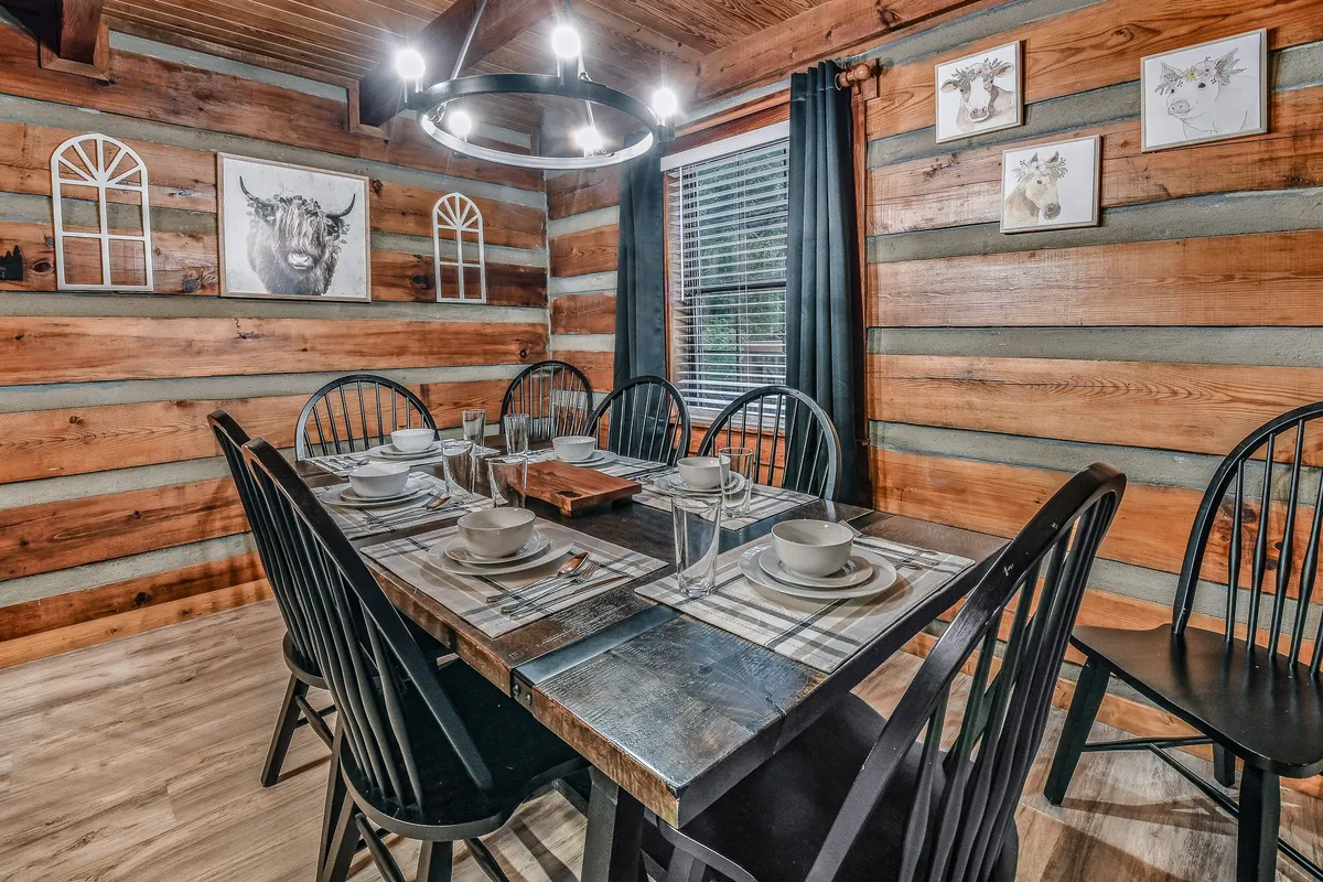 A large wooden dining table surrounded by black chairs stands ready for group meals or game nights. The décor is rustic, with decorative wall art and wooden walls enhancing the mountain cabin vibe. The spaciousness and setting invite shared experiences and celebrations.
