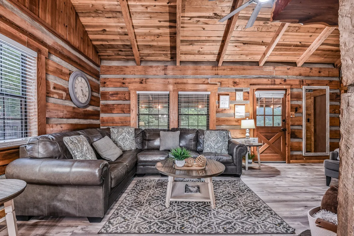 A comfortable living room with a leather sectional sofa, decorative pillows, and a coffee table with plants. Wooden beams and a large wall clock add rustic charm, while the open arrangement is perfect for entertaining or relaxing with loved ones.