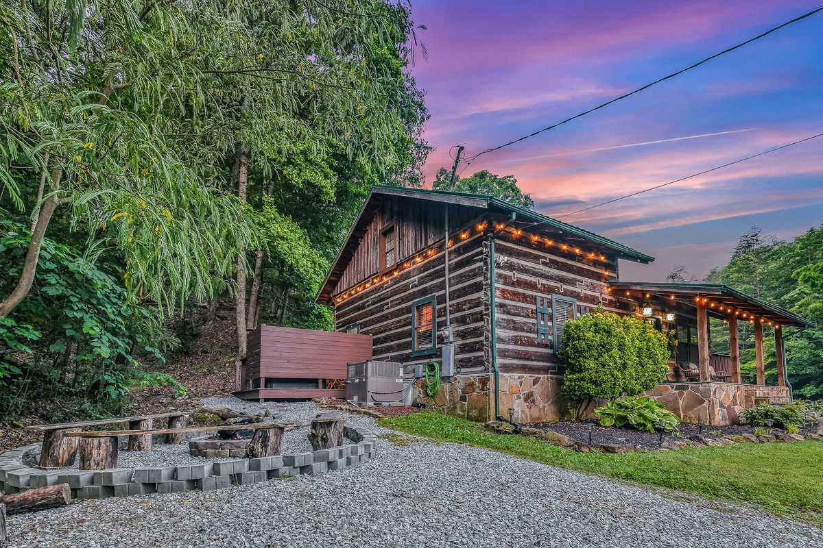 A charming log cabin is seen illuminated by twinkling exterior lights, with a well-arranged fire pit area and lush greenery nearby. The outdoor space invites gatherings, featuring comfortable seating around the fire—ideal for stories and s’mores.