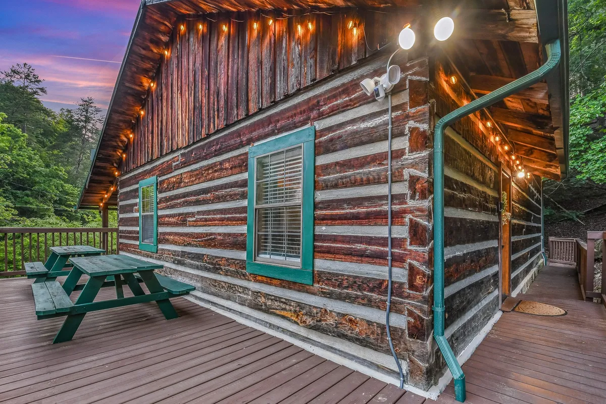 This image features the exterior of a log cabin with green-trimmed windows and an inviting outdoor seating area. The surrounding trees and natural landscape create a sense of connection to the Smoky Mountains, inviting families to enjoy outdoor dining, evening gatherings, or stargazing from the deck.