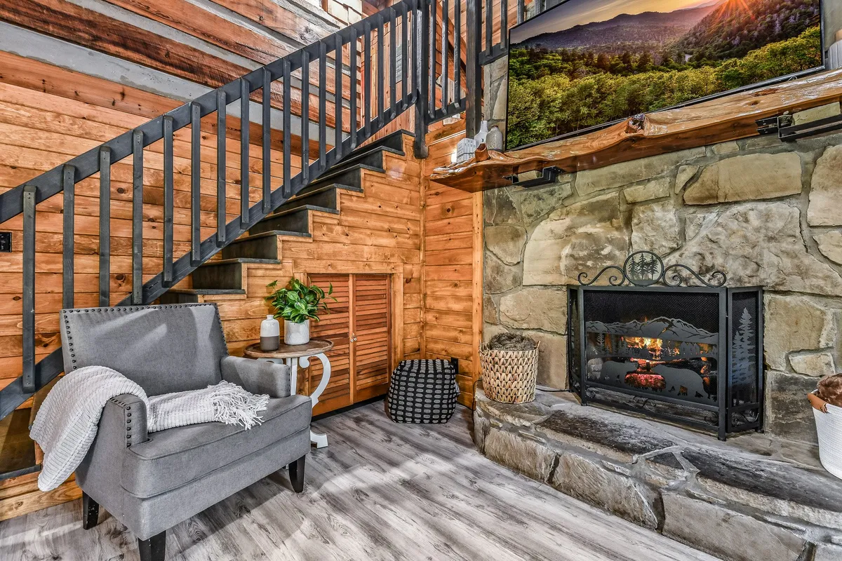 A warm, welcoming living area featuring an electric fireplace with a cozy armchair nearby, a staircase leading up, and a television positioned for easy viewing. The space is styled with wooden finishes and ambient lighting, creating an inviting setting for relaxation or socializing.