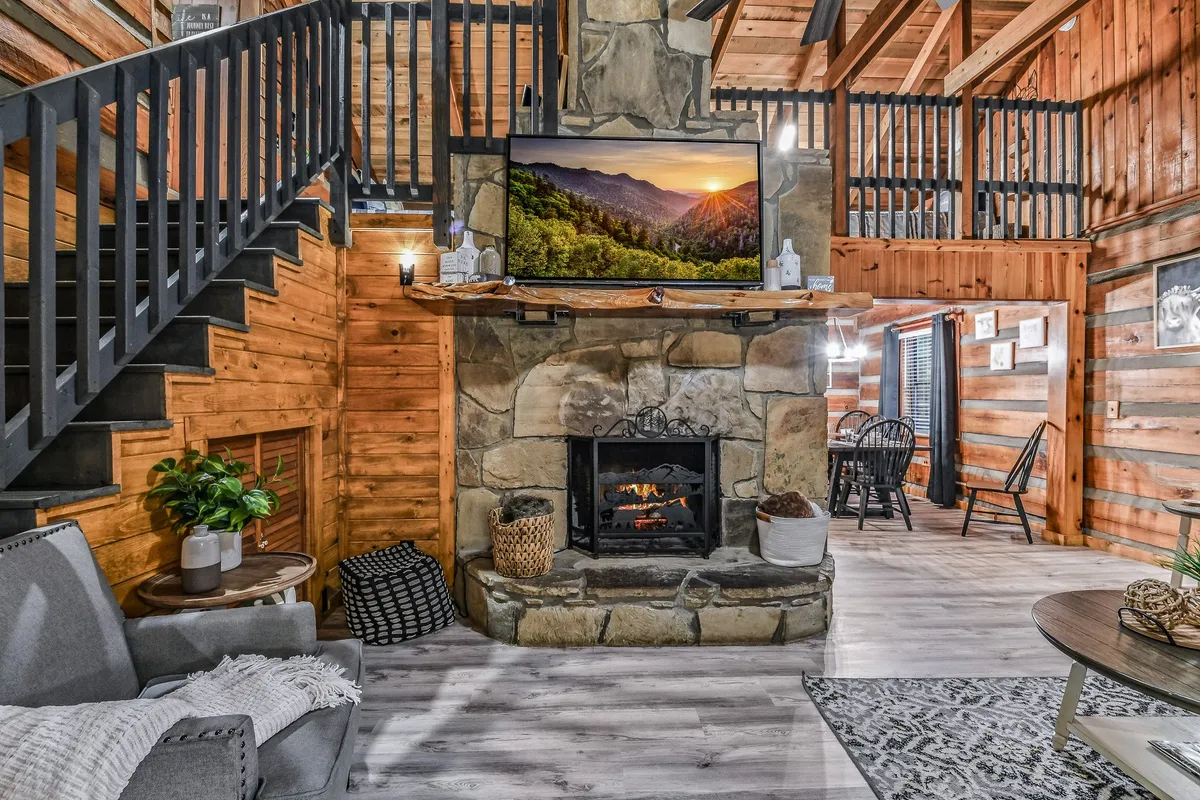This inviting living area features a stone fireplace as the focal point, a large TV above, and comfortable seating. The wooden staircase and open dining area in the background hint at the home's spacious layout. Natural light brightens the space, suggesting a warm atmosphere for gatherings, movie nights, or winding down after a day out.