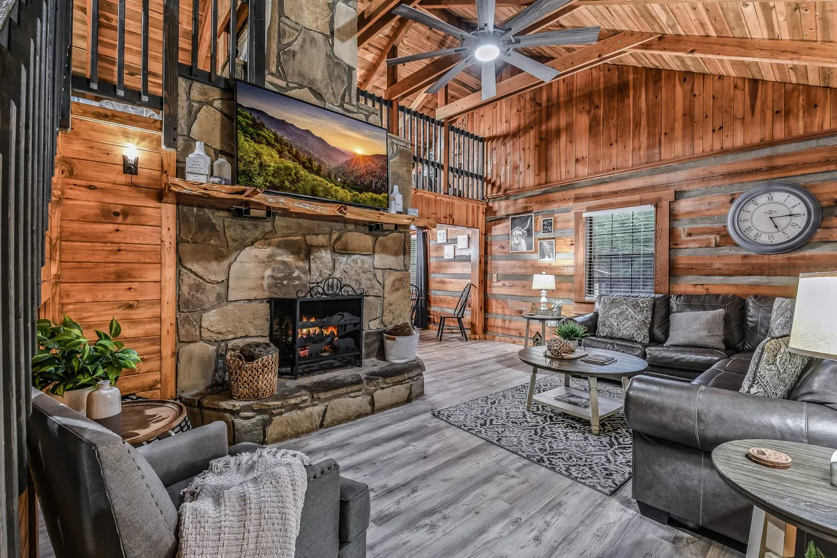 A warm, spacious living area featuring a large stone fireplace, comfortable seating, wooden beams, and a TV displaying a mountain vista. The setting is designed for relaxation, socializing, and spending quality time with loved ones—including pets.